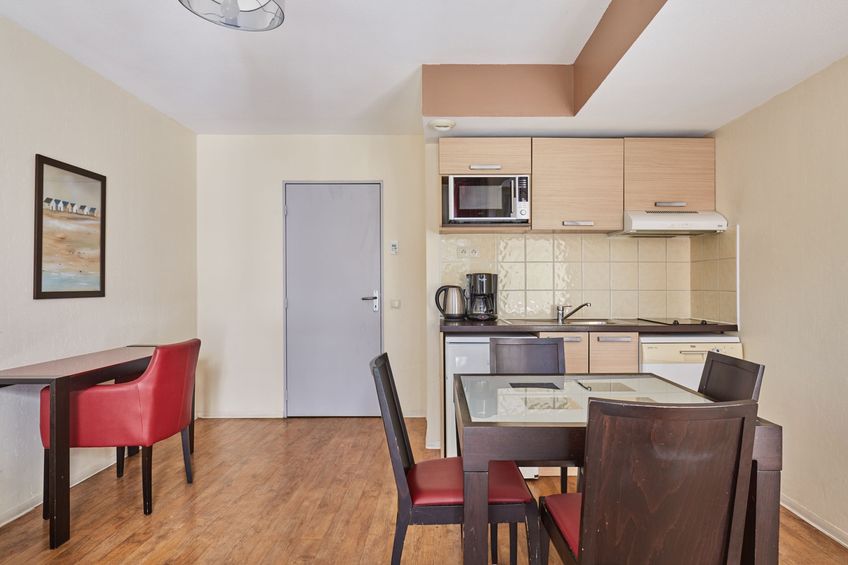 A compact kitchen with a microwave, coffee maker, and a dishwasher. There is a dining table with chairs and a framed picture on the wall.