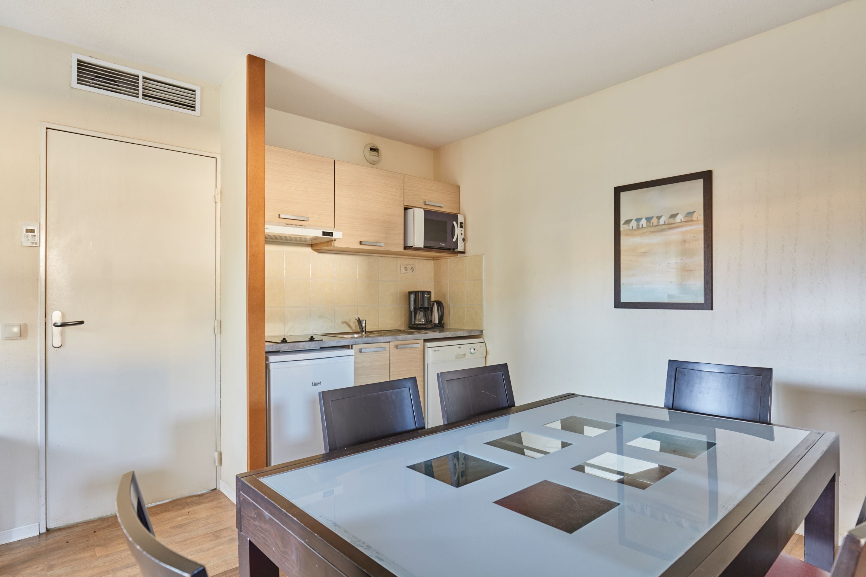 Una cocina compacta equipada con microondas, cafetera y armarios. Se aprecia una mesa de comedor con sillas y un cuadro en la pared.