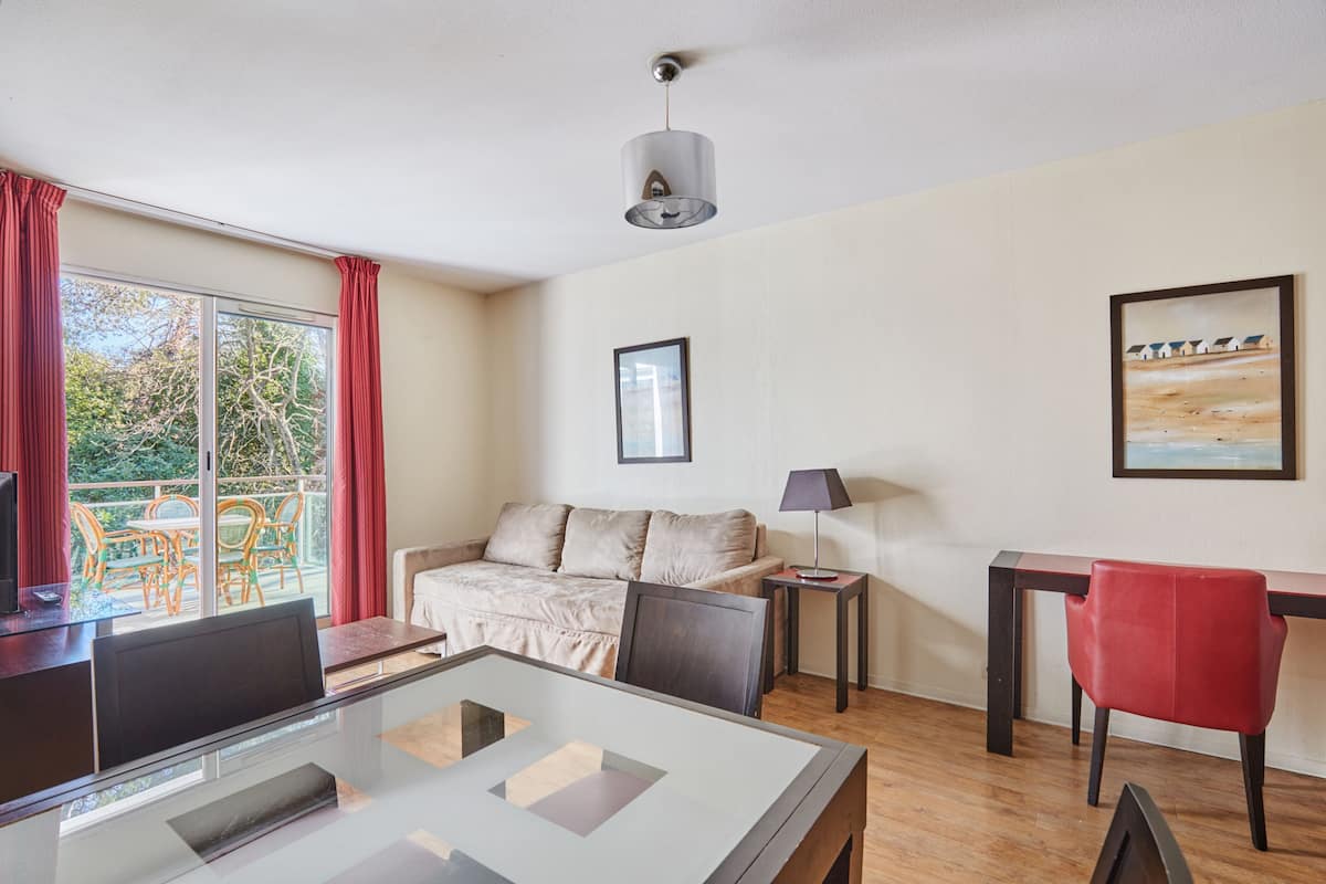 Ein Zimmer mit Glastisch, einer Couch, einem roten Sessel, einer Lampe und einem Gemälde an der Wand.