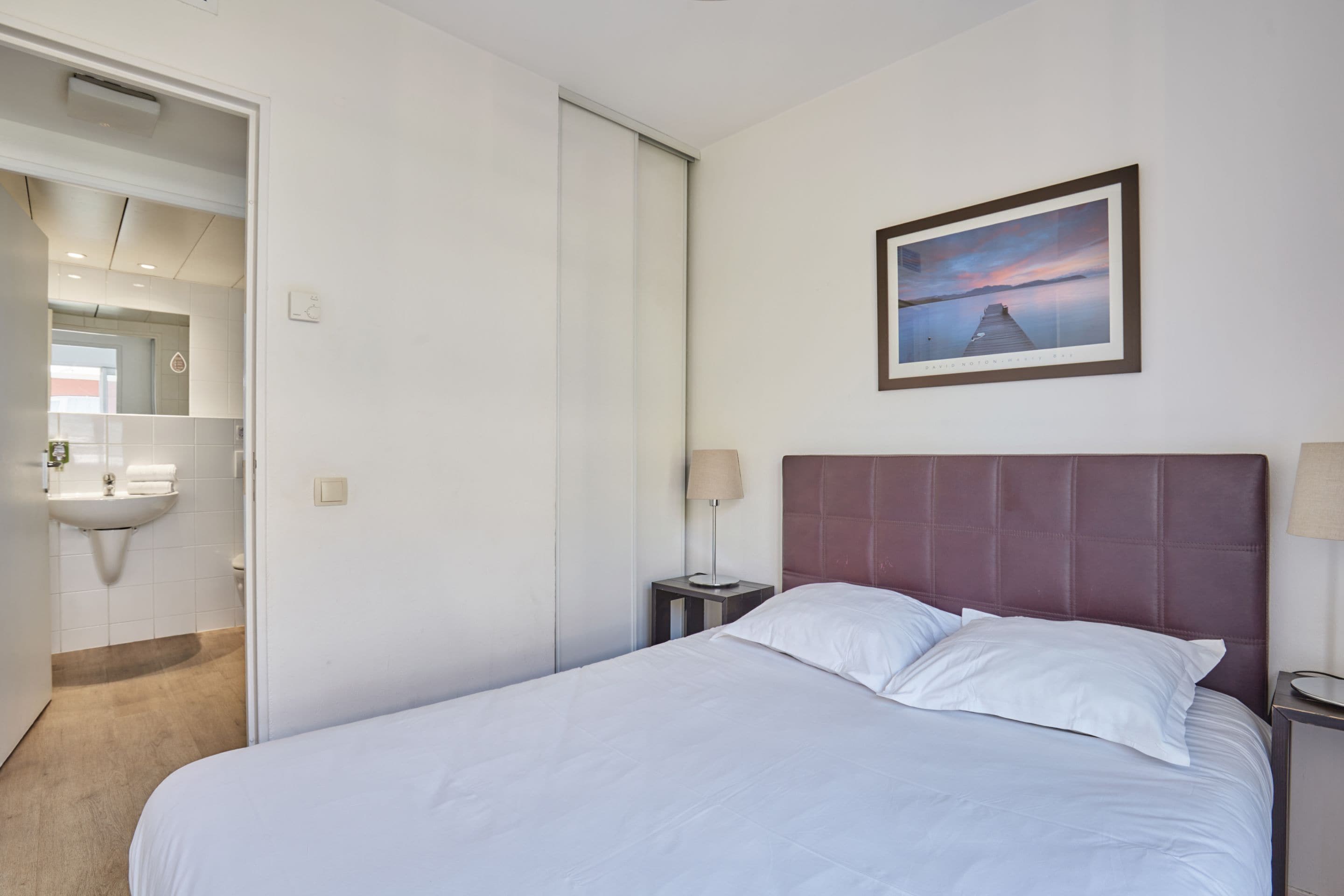 A hotel room with a bed, a nightstand, a lamp, a framed picture on the wall, and a bathroom visible through an open door.