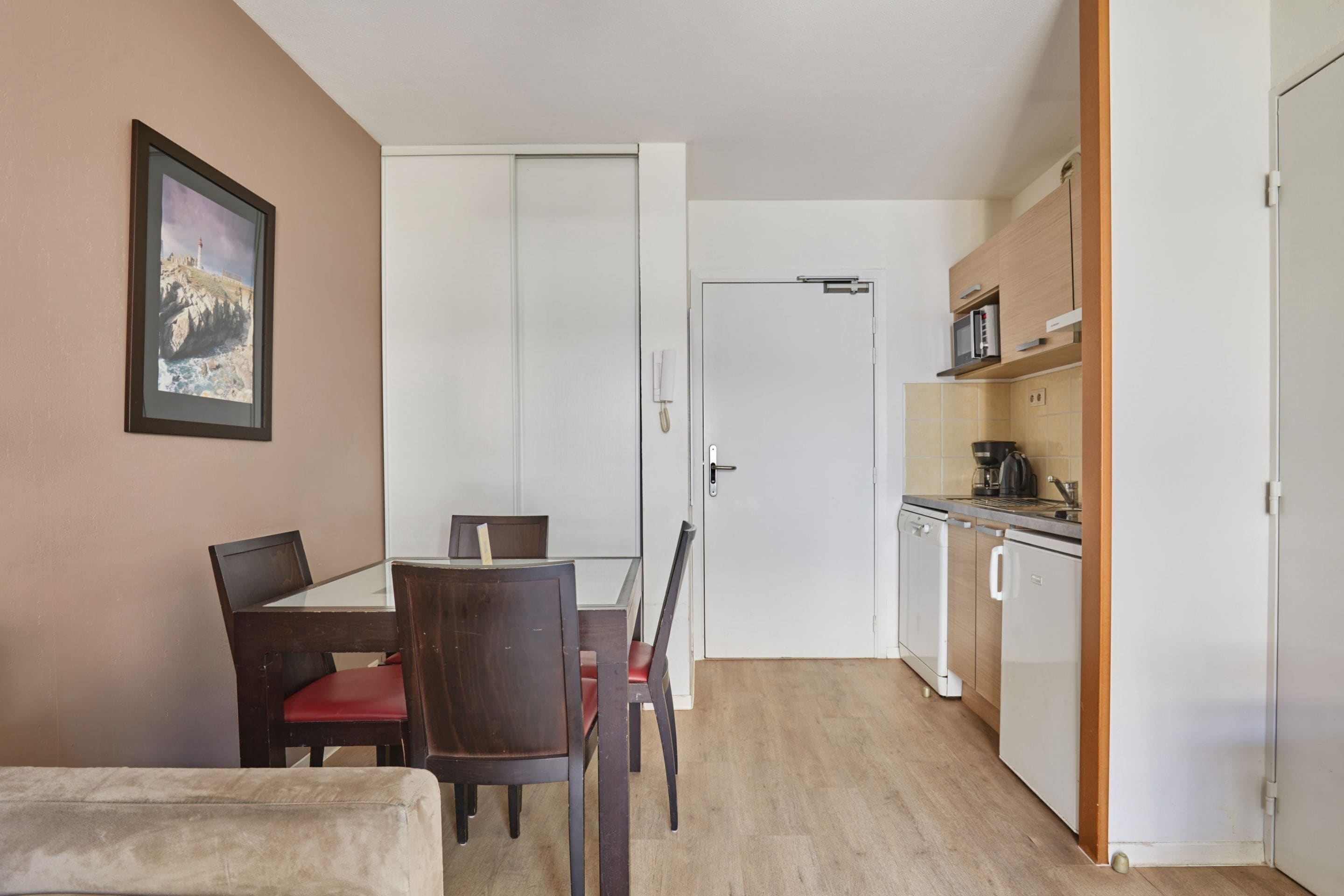 Een kleine keuken met een magnetron, kookplaat en keukenkastjes, een eethoek met een tafel en stoelen, en een lijst met een afbeelding aan de muur.