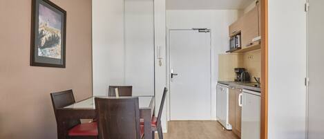 Een kleine keuken met een magnetron, kookplaat en keukenkastjes, een eethoek met een tafel en stoelen, en een lijst met een afbeelding aan de muur.