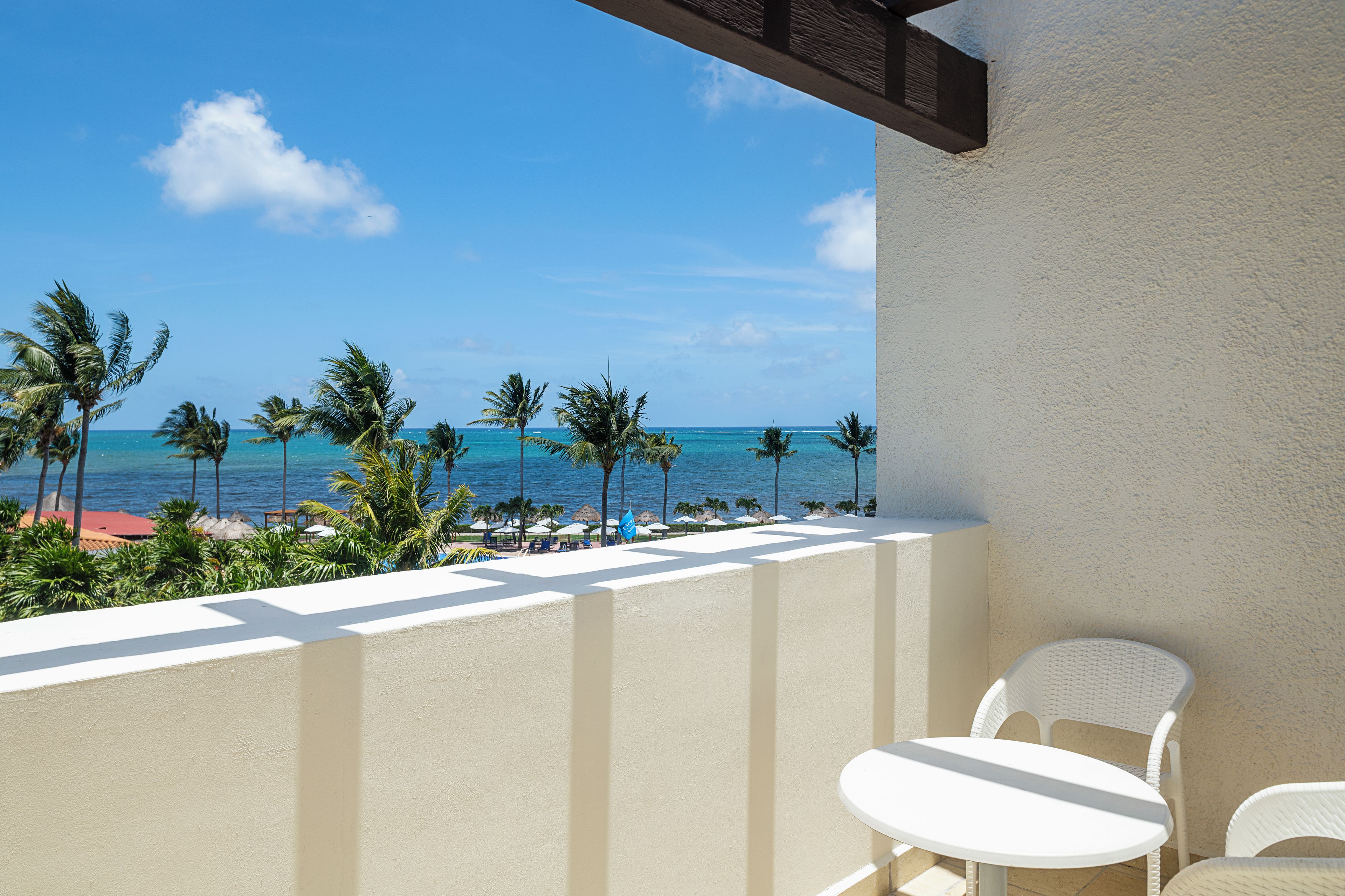 A balcony with a view of the ocean, palm trees, and a white table and chair.