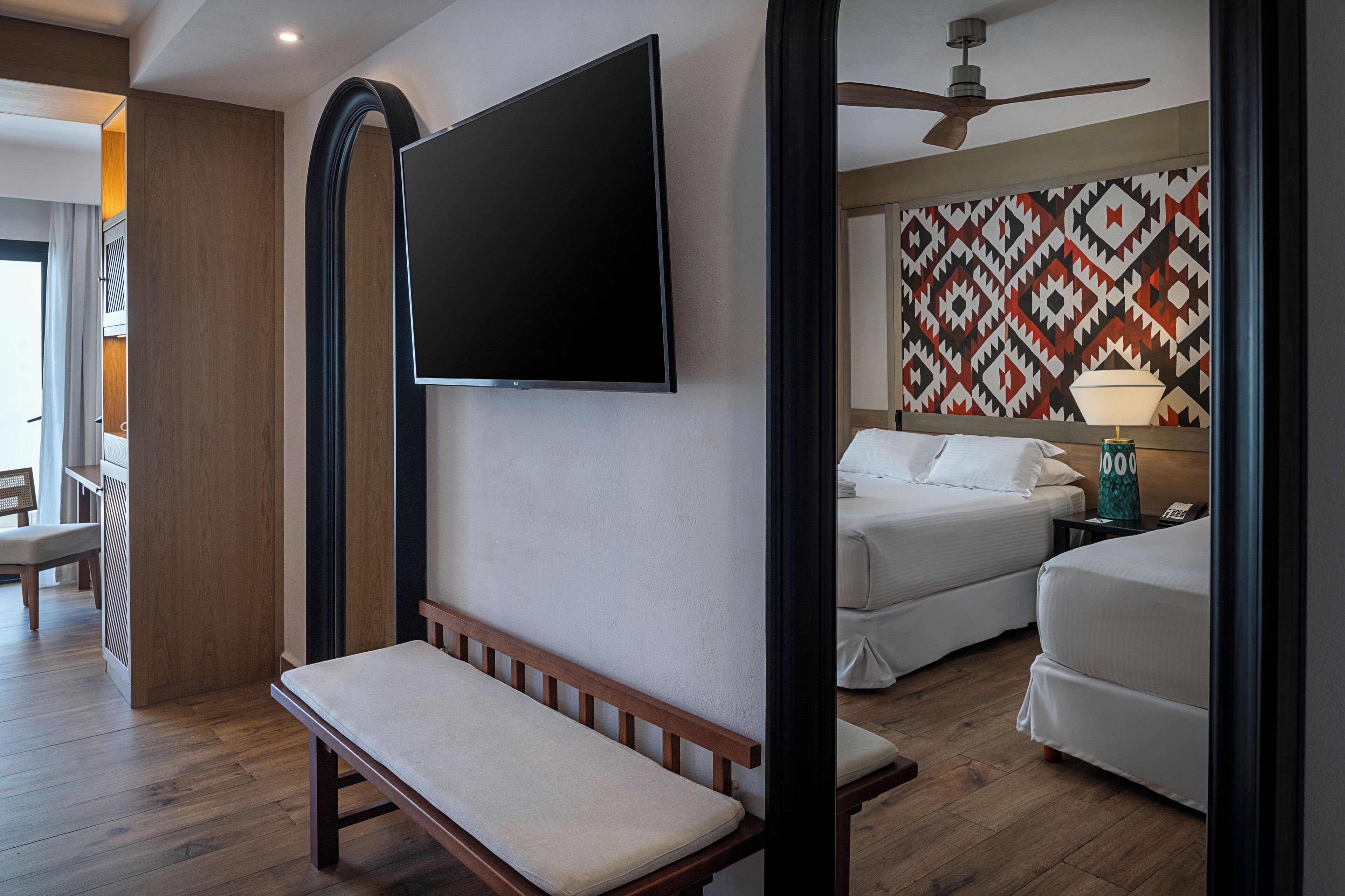 Hotelkamer met een wandgemonteerde flatscreen televisie, een houten bankje, twee bedden, een plafondventilator en een behang met patroon.