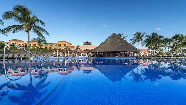 A clear blue pool with lounge chairs and a thatched-roof structure, surrounded by palm trees and resort buildings.