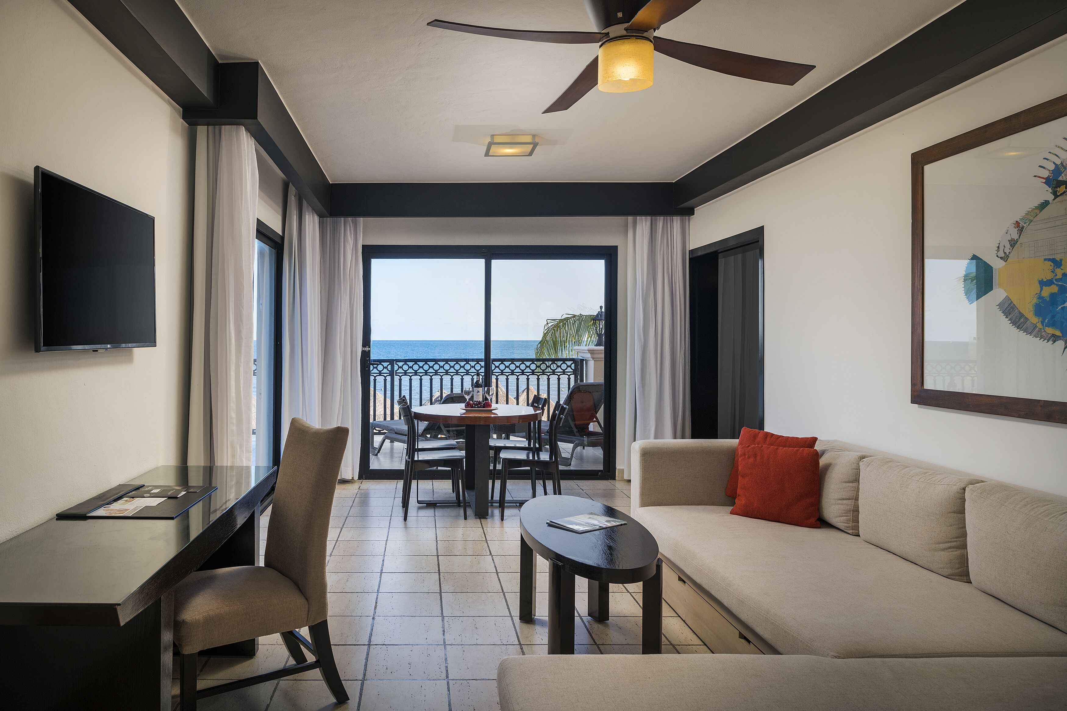 A hotel room with a balcony, a dining table, a sofa, a TV, and a ceiling fan.