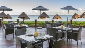 An outdoor dining area with tables and chairs, thatched umbrellas, and a view of the ocean.