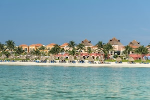 A beachfront hotel with thatched-roof buildings, palm trees, and a row of beachside chairs.