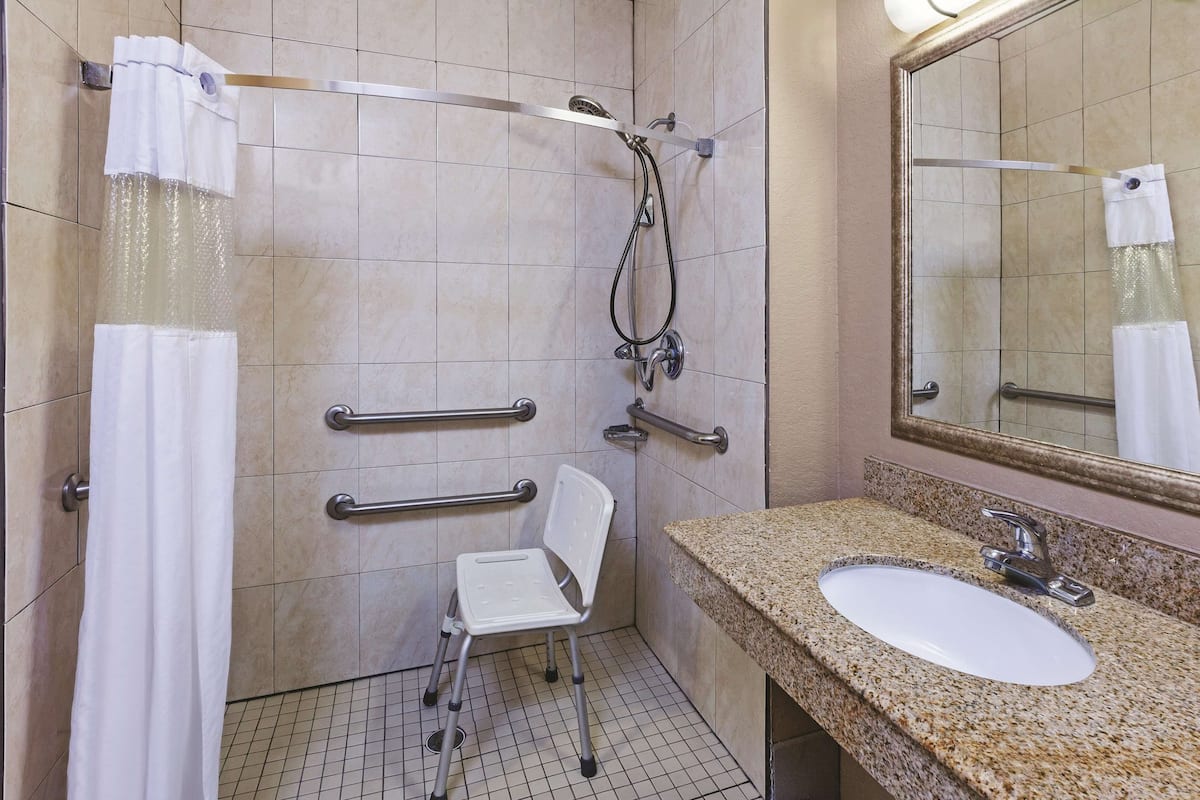 A bathroom with a shower, grab bars, a wheelchair-accessible chair, a sink, and a mirror.