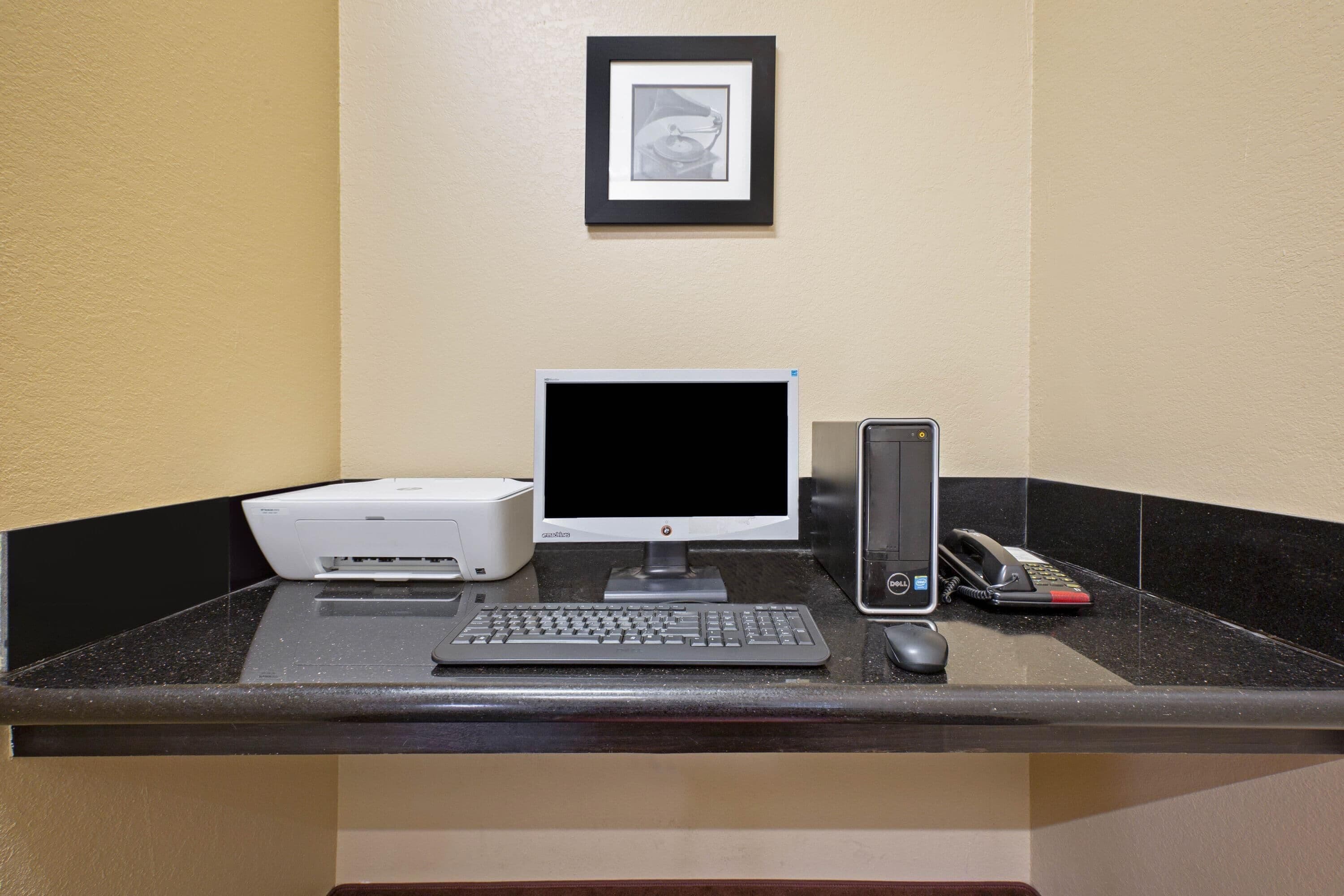 Un comptoir d’accueil d’hôtel avec un ordinateur de bureau, une imprimante, un téléphone et un tableau encadré accroché au mur.