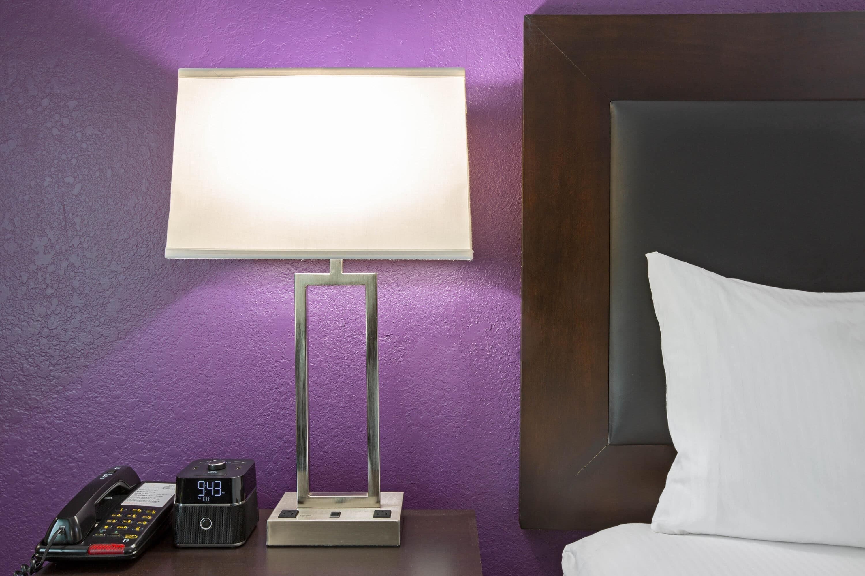 A hotel room with a bed, a nightstand with a lamp, a telephone, and a digital clock.