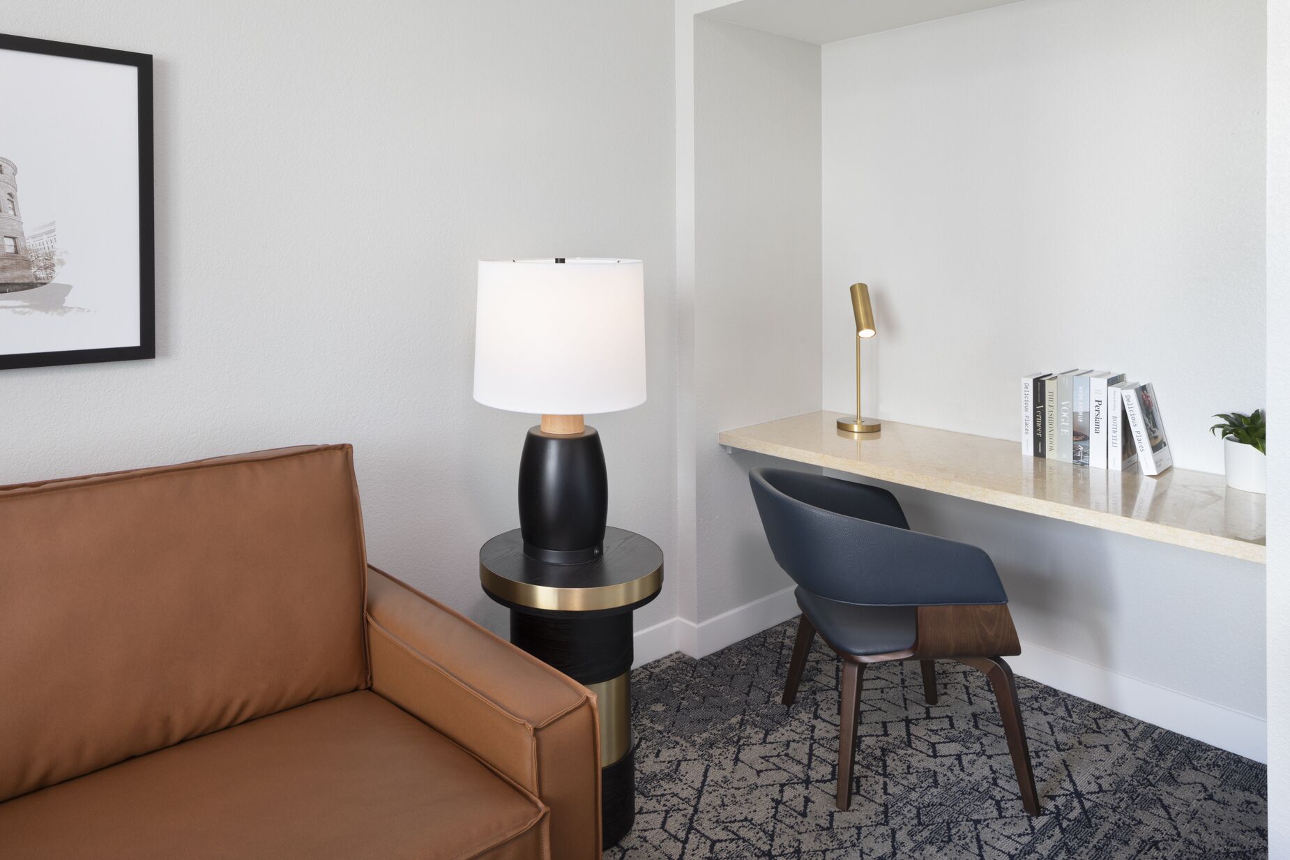 A hotel room with a brown leather sofa, a round black and gold side table with a white lamp, a desk with a gold lamp, and a chair. There is a framed picture on the wall and a small potted plant on the desk.