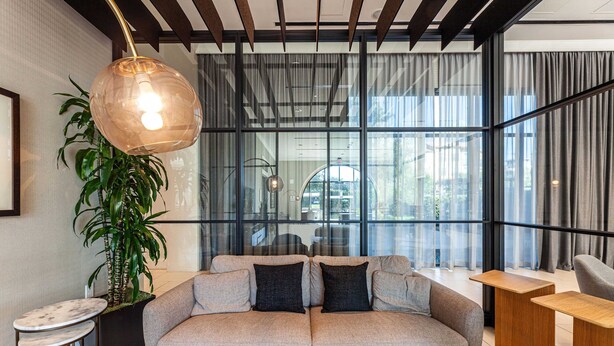 A modern living room with a sofa, a hanging light fixture, a potted plant, and a view through large windows. - Sheraton Stonebriar Hotel (Frisco)