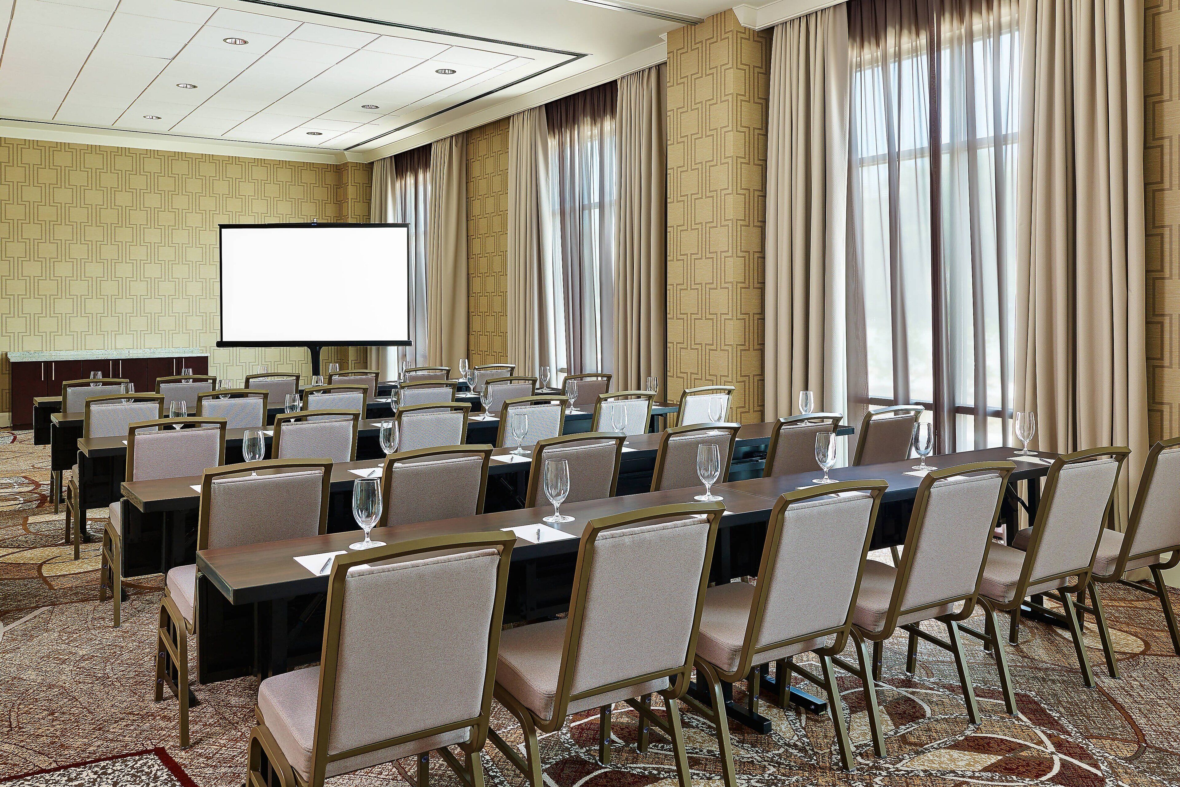 Un salón de conferencias con varias mesas y sillas, una pantalla de proyector y ventanas amplias con cortinas.