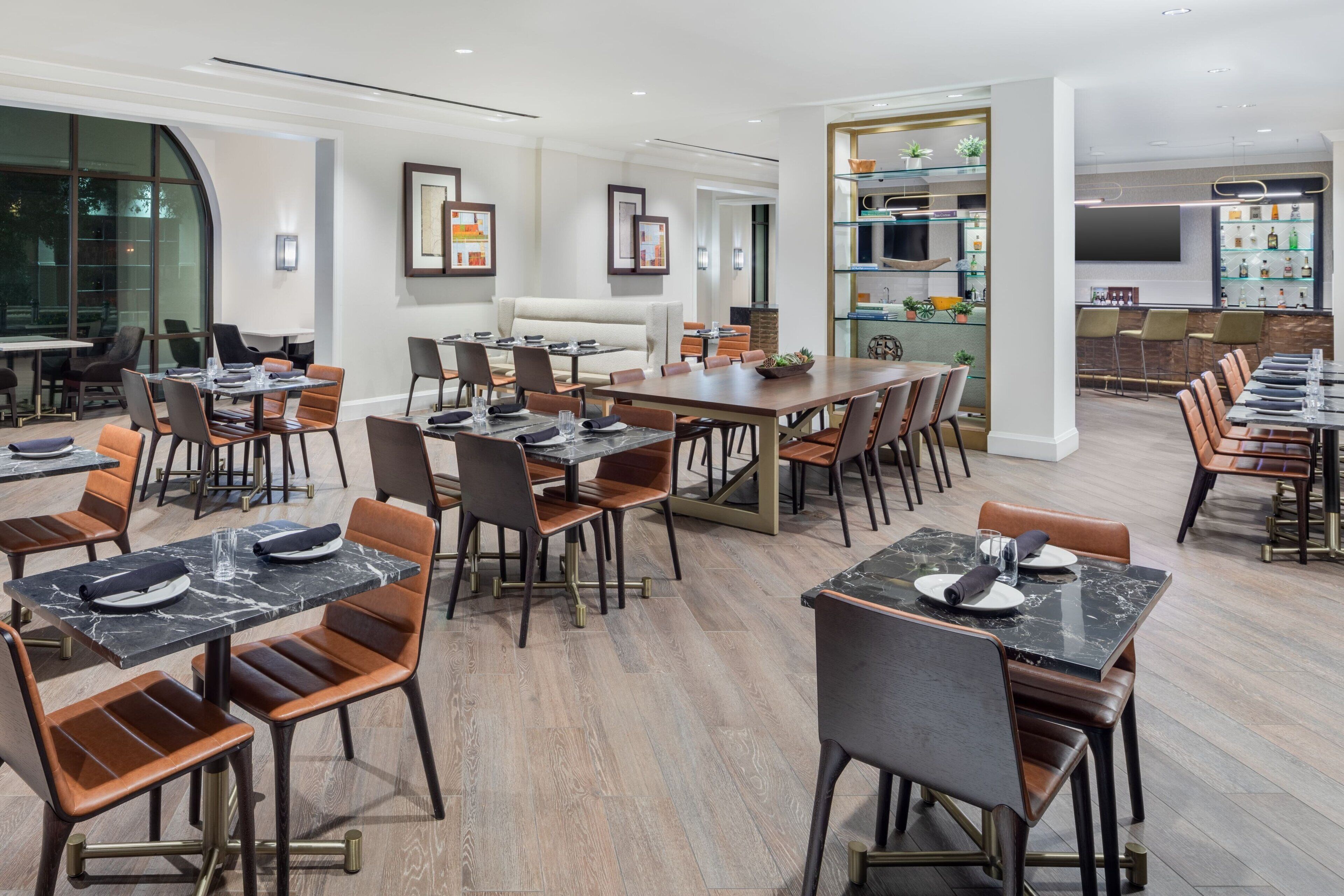 Un comedor moderno de hotel con varias mesas y sillas, una zona de bar con estanterías, y un amplio ventanal que ofrece vistas al exterior.