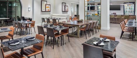 Un comedor moderno de hotel con varias mesas y sillas, una zona de bar con estanterías, y un amplio ventanal que ofrece vistas al exterior.