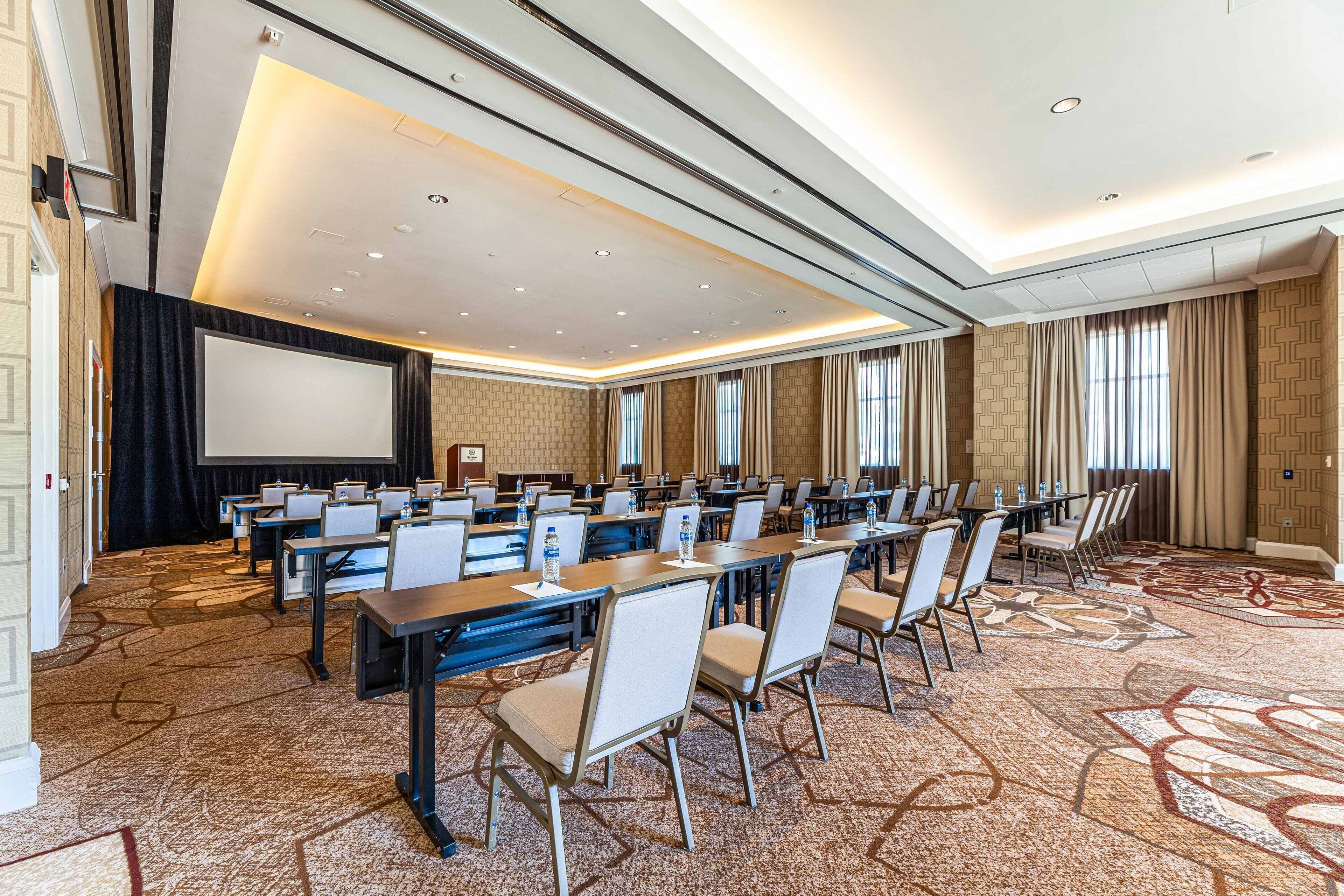A conference room with rows of tables and chairs, a projector screen, and a carpeted floor.