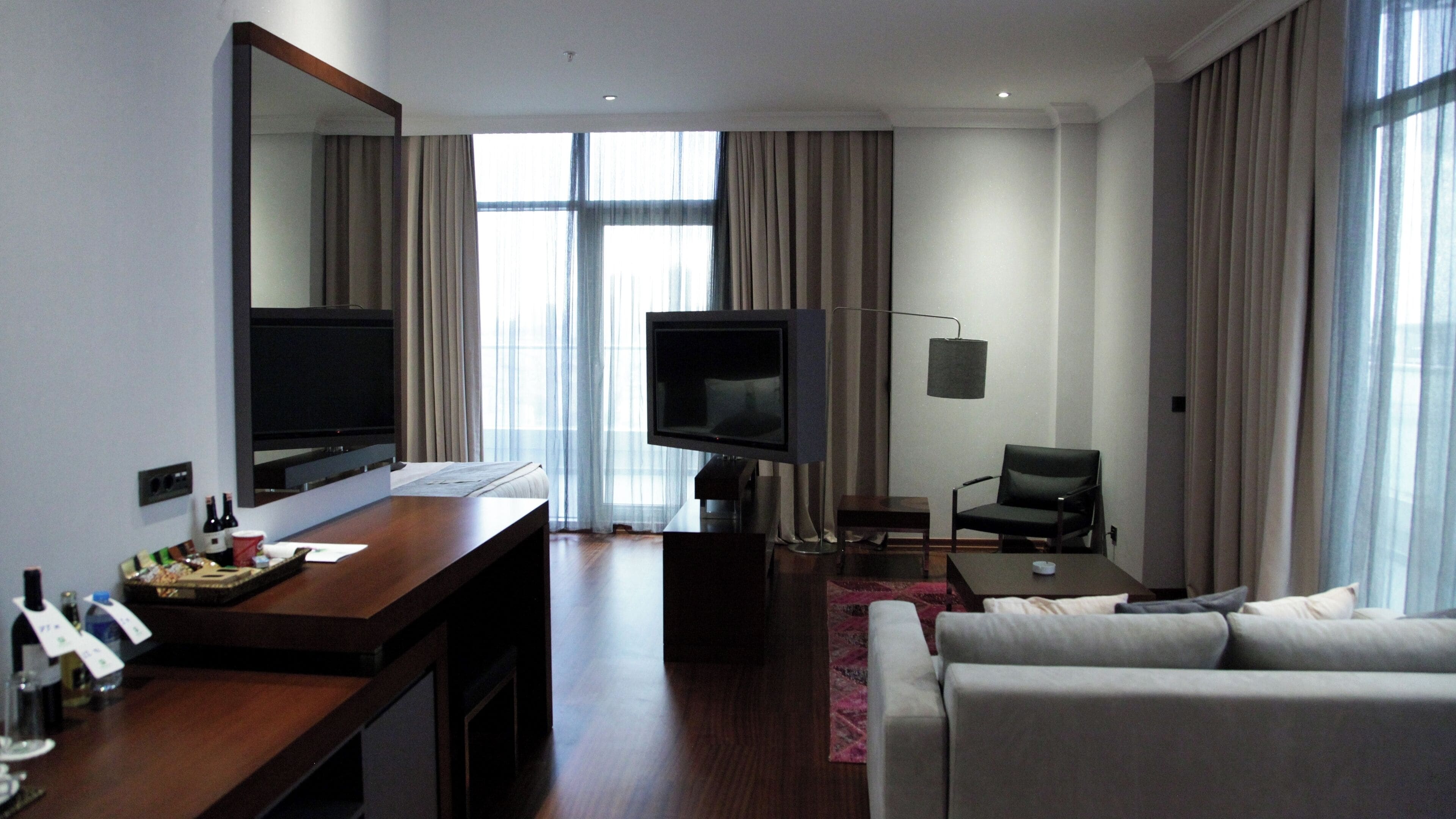 A modern hotel room with a large flat-screen TV, a sofa, a dining table, and a large mirror.