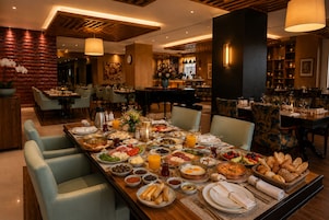 Un área de comedor con una mesa larga dispuesta para una comida, rodeada de sillas, con una barra y un piano al fondo.