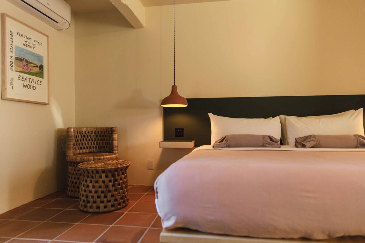 A neatly arranged bedroom with a bed, pillows, a hanging light fixture, a wicker chair, and a small table.