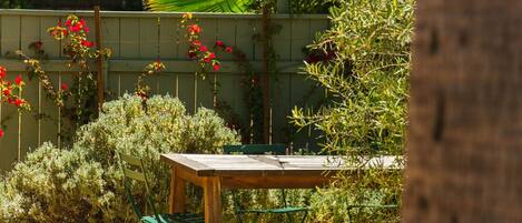Un conjunto de mesa y banco de madera en un jardín, con una valla de madera, rodeado de diversas plantas y una guirnalda de luces colgantes.