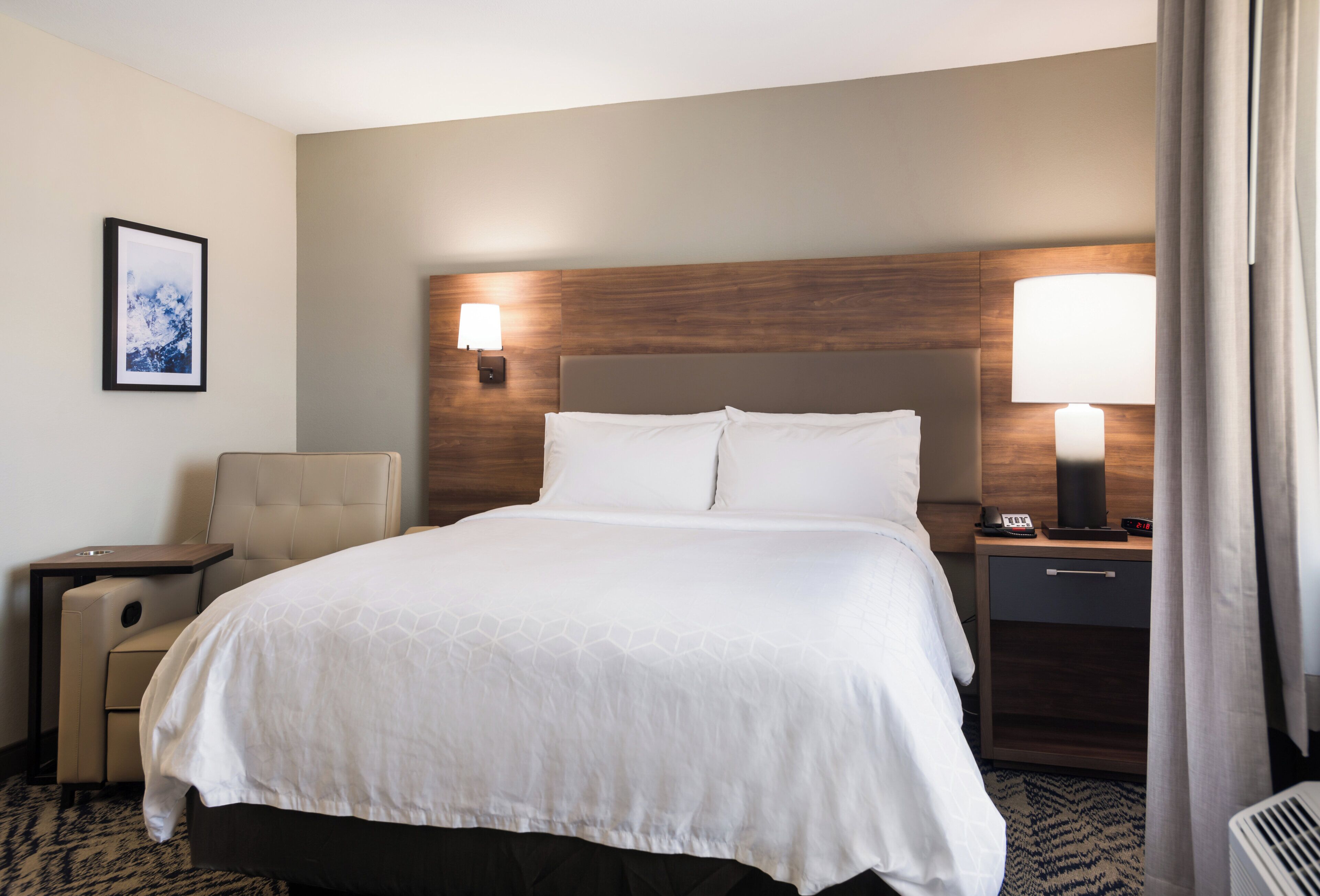 A hotel room with a large bed, a chair, a desk, a nightstand with a lamp, and a framed picture on the wall.