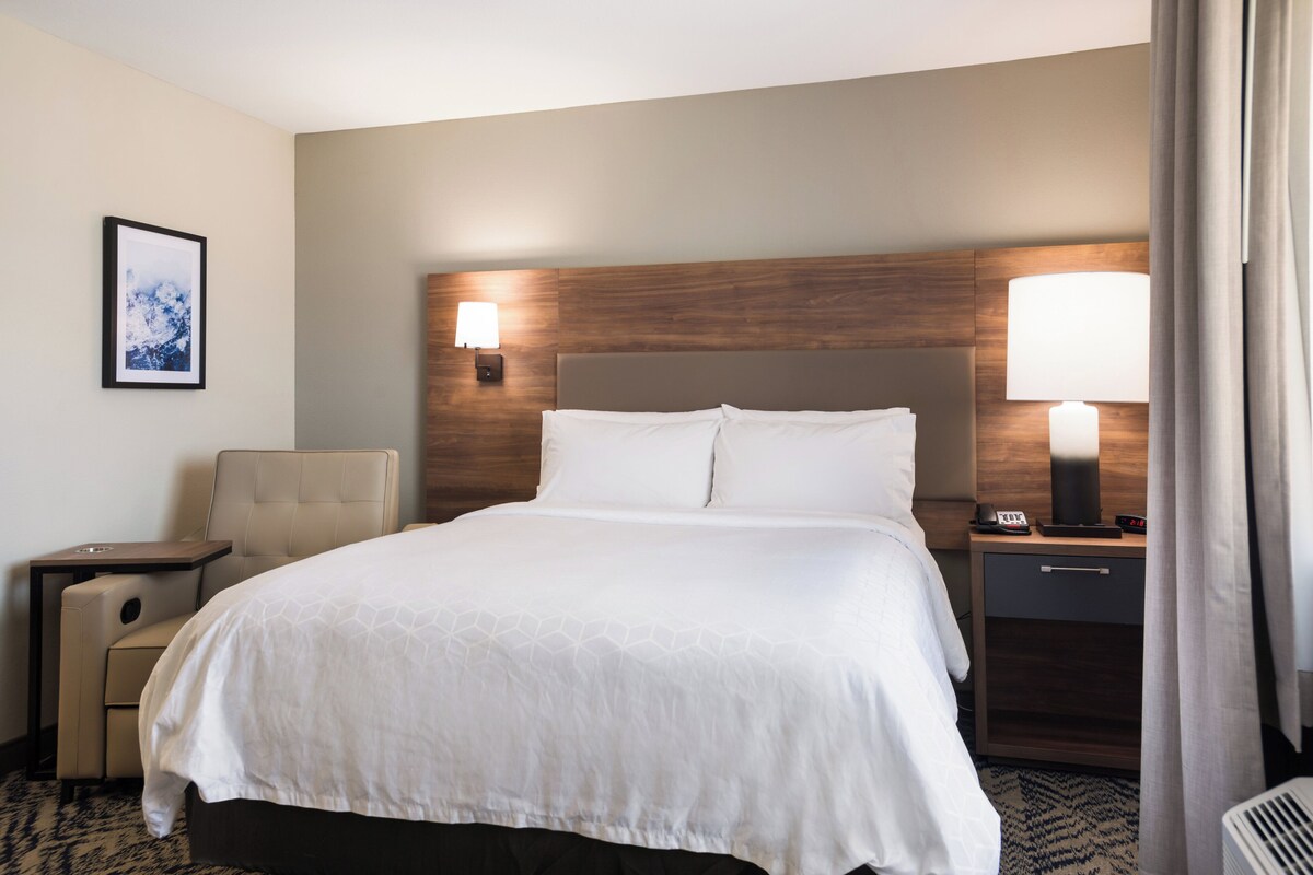 Ein Hotelzimmer mit einem großen Bett, einem Sessel, einem Schreibtisch, einem Nachttisch mit Lampe und einem gerahmten Bild an der Wand.
