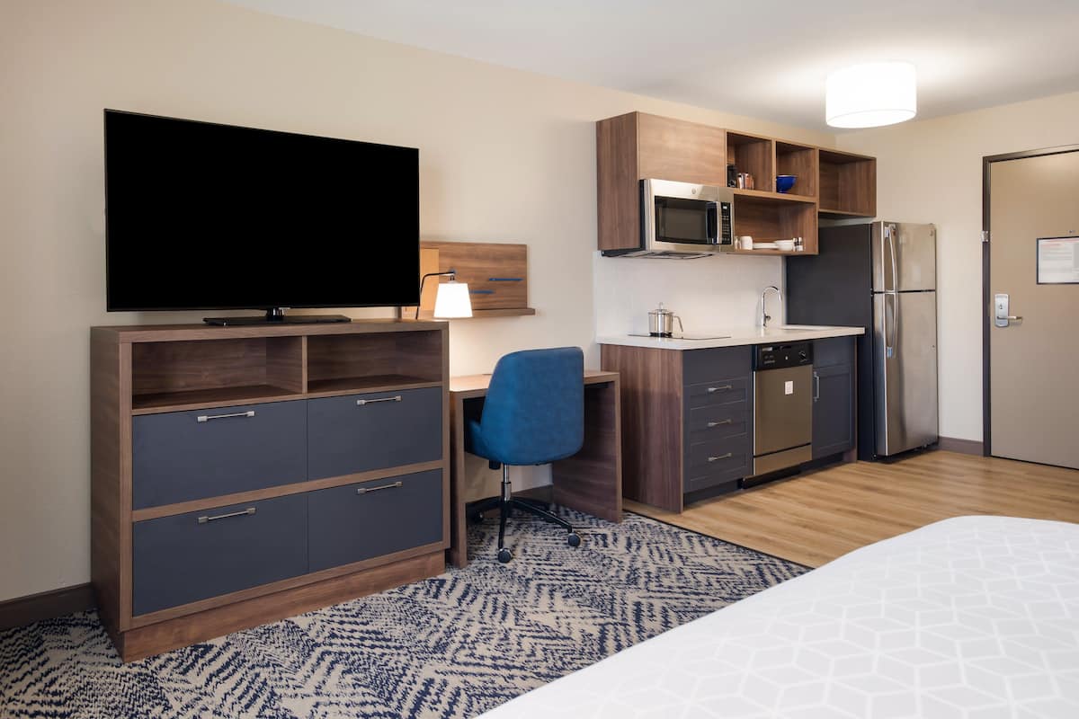 Ein Hotelzimmer mit einem Bett, einem Schreibtisch mit Stuhl, einem Fernseher, einer Küchenzeile mit Mikrowelle und Kühlschrank sowie einem gemusterten Teppich.