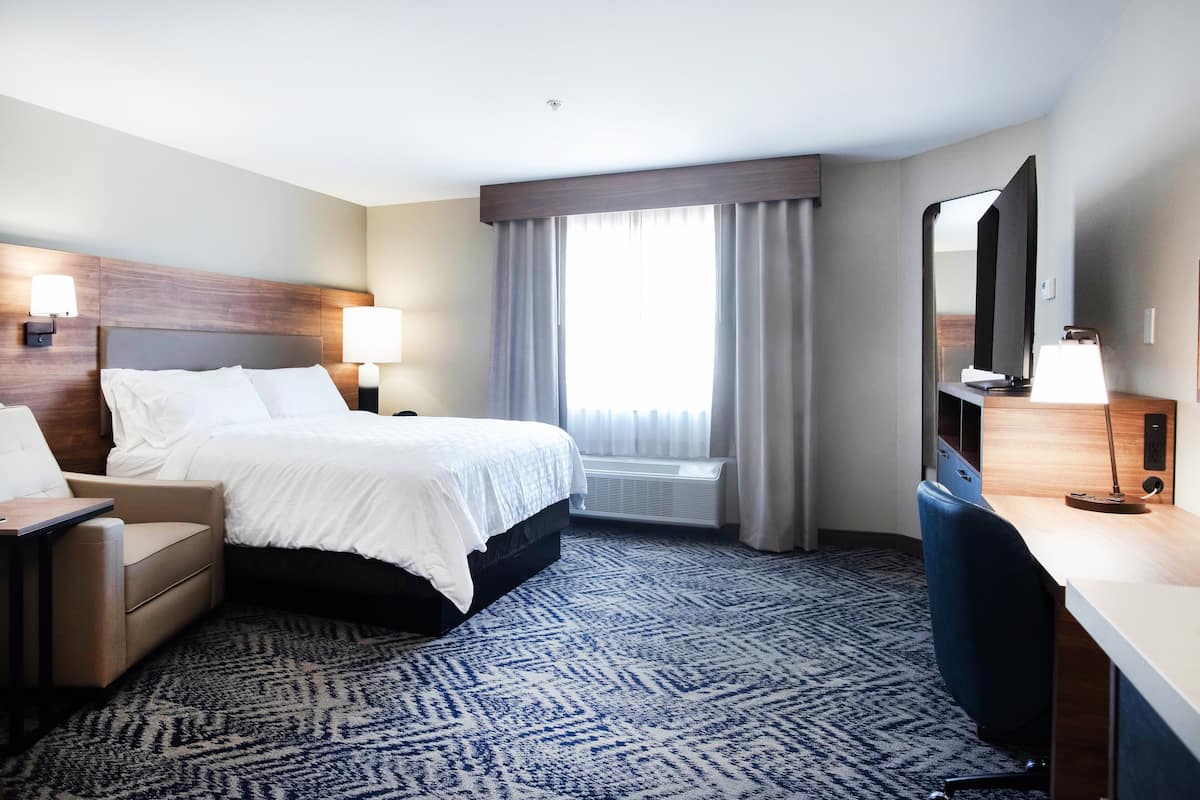 Ein Hotelzimmer mit Bett, Schreibtisch, Stuhl, Schreibtischlampe, Nachttisch und einem Fenster mit Vorhängen.