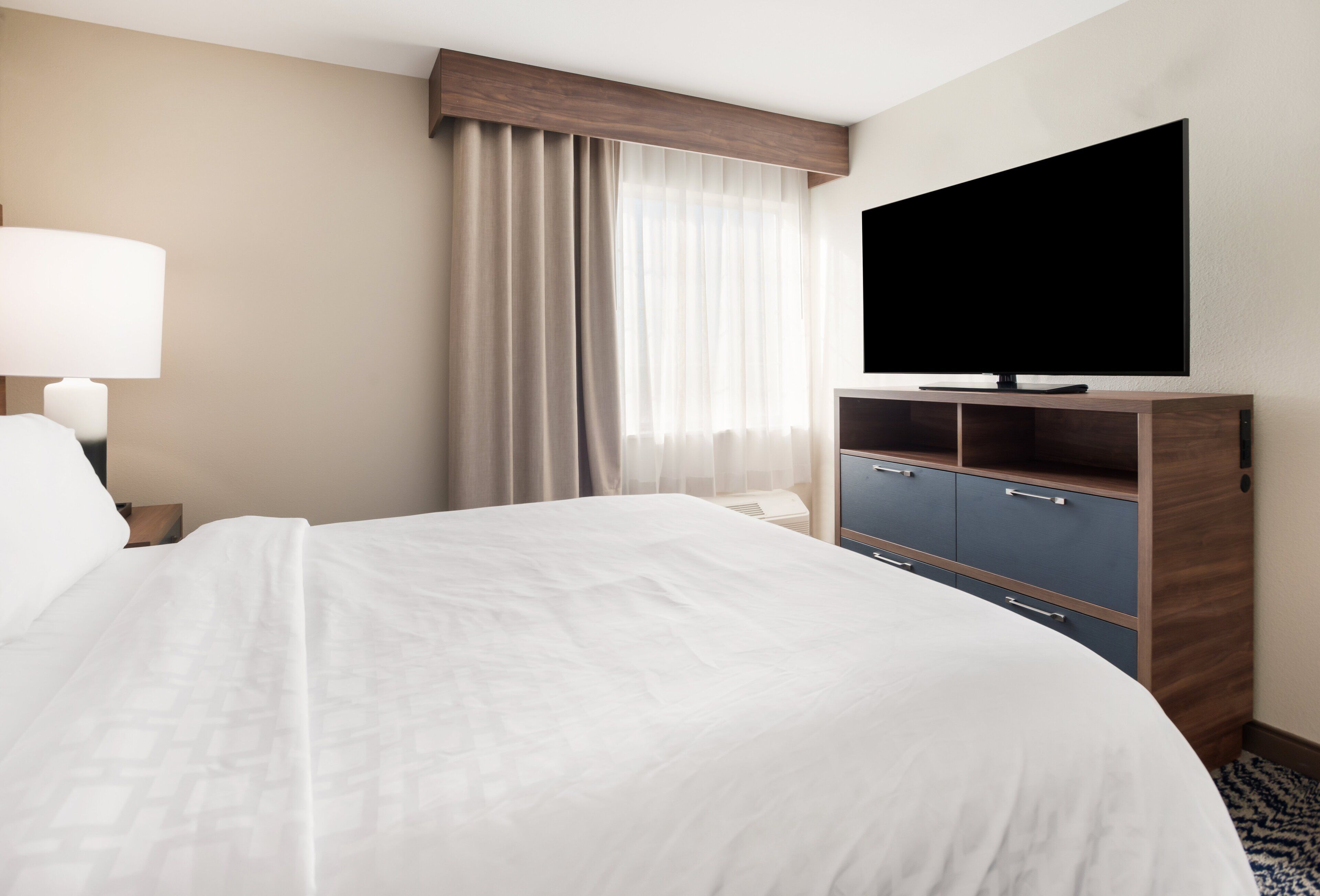 Habitación de hotel con una cama grande, un televisor de pantalla plana, un escritorio con lámpara, un armario y una ventana con cortinas.
