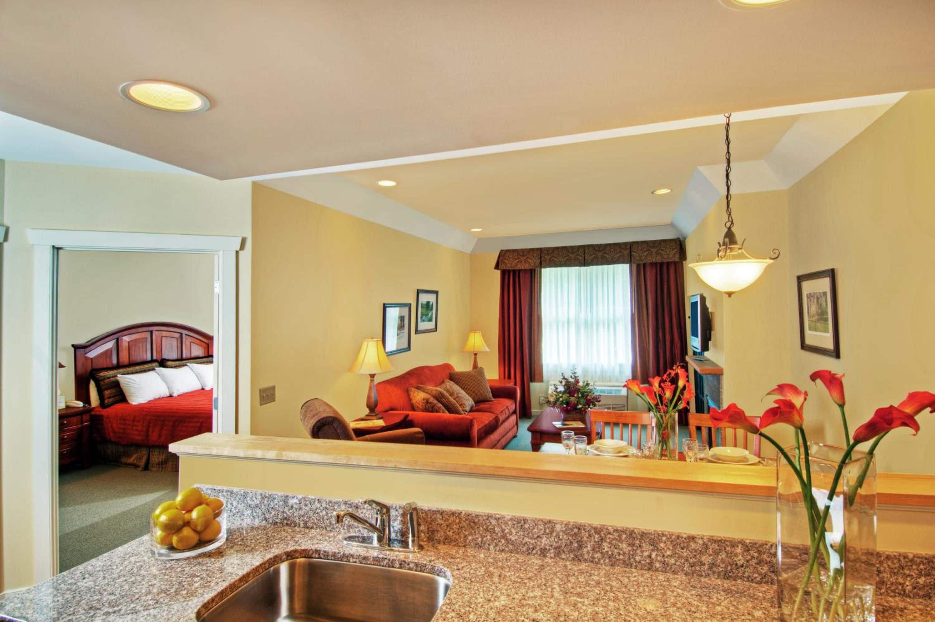 Habitación de hotel con área de cocina, una mesa de comedor, un sofá rojo y una cama con cabecera roja.