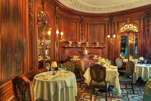 Un comedor formal con paneles de madera ornamentados, varias mesas preparadas para el servicio y un espejo que refleja la decoración del salón.