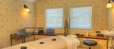 A room with two massage tables, a wooden bench with towels, a shelf with toiletries, and a window with blinds.