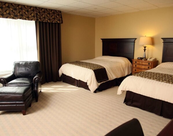 A hotel room with two beds, a leather armchair, a bedside table with a lamp, and a window with curtains.