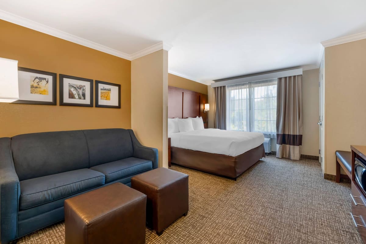 A hotel room with a blue sofa, a bed with white linens, a small table, and a large window with curtains.