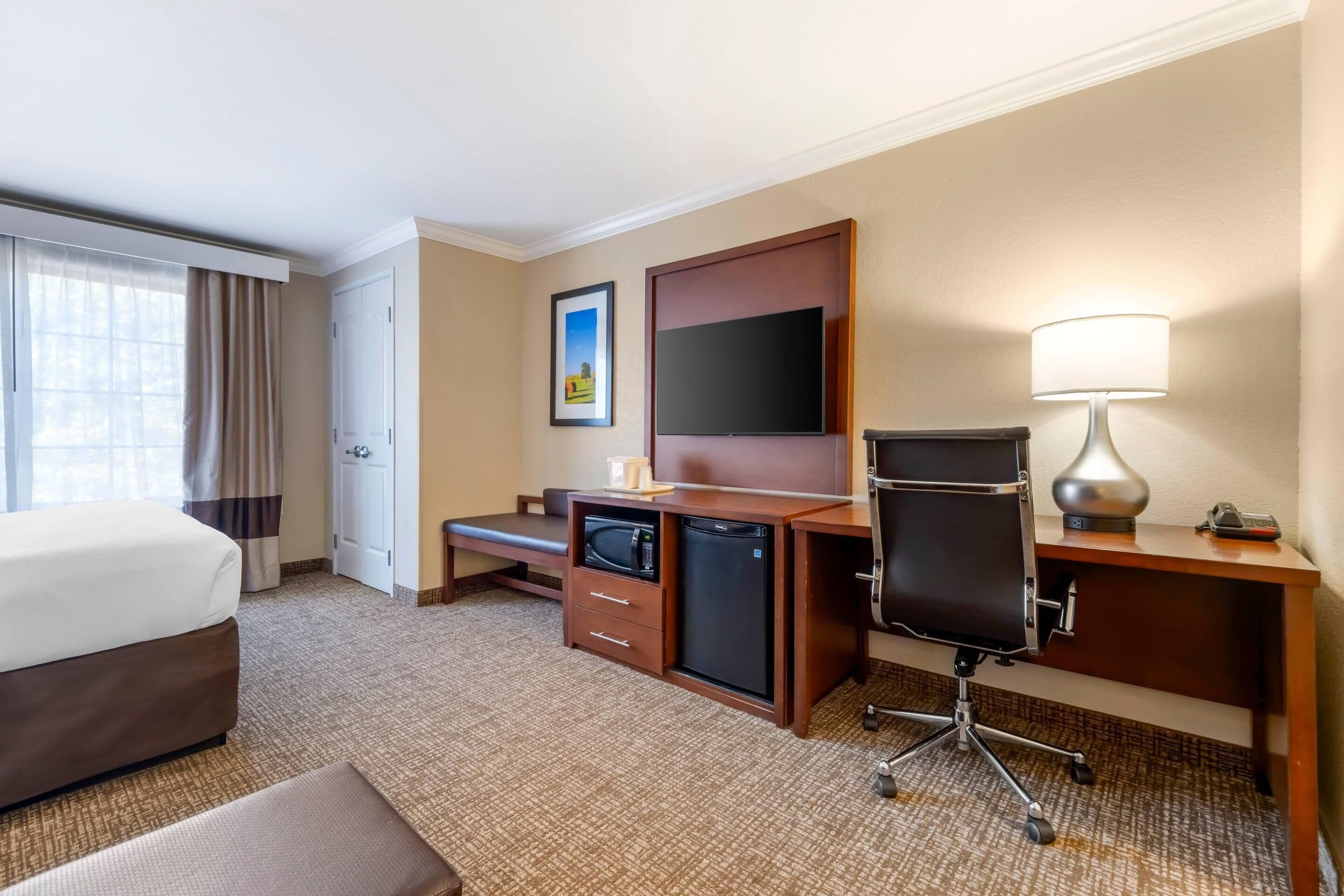 A hotel room with a bed, a desk with a computer, a television, a lamp, and a small refrigerator.