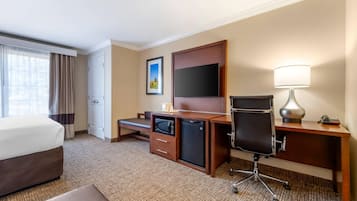 A hotel room with a bed, a desk with a computer, a television, a lamp, and a small refrigerator.