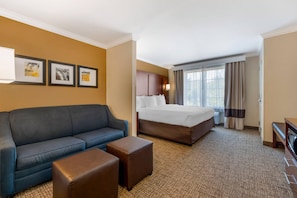 A hotel room with a blue sofa, a bed with white linens, a small table, and a large window with curtains.