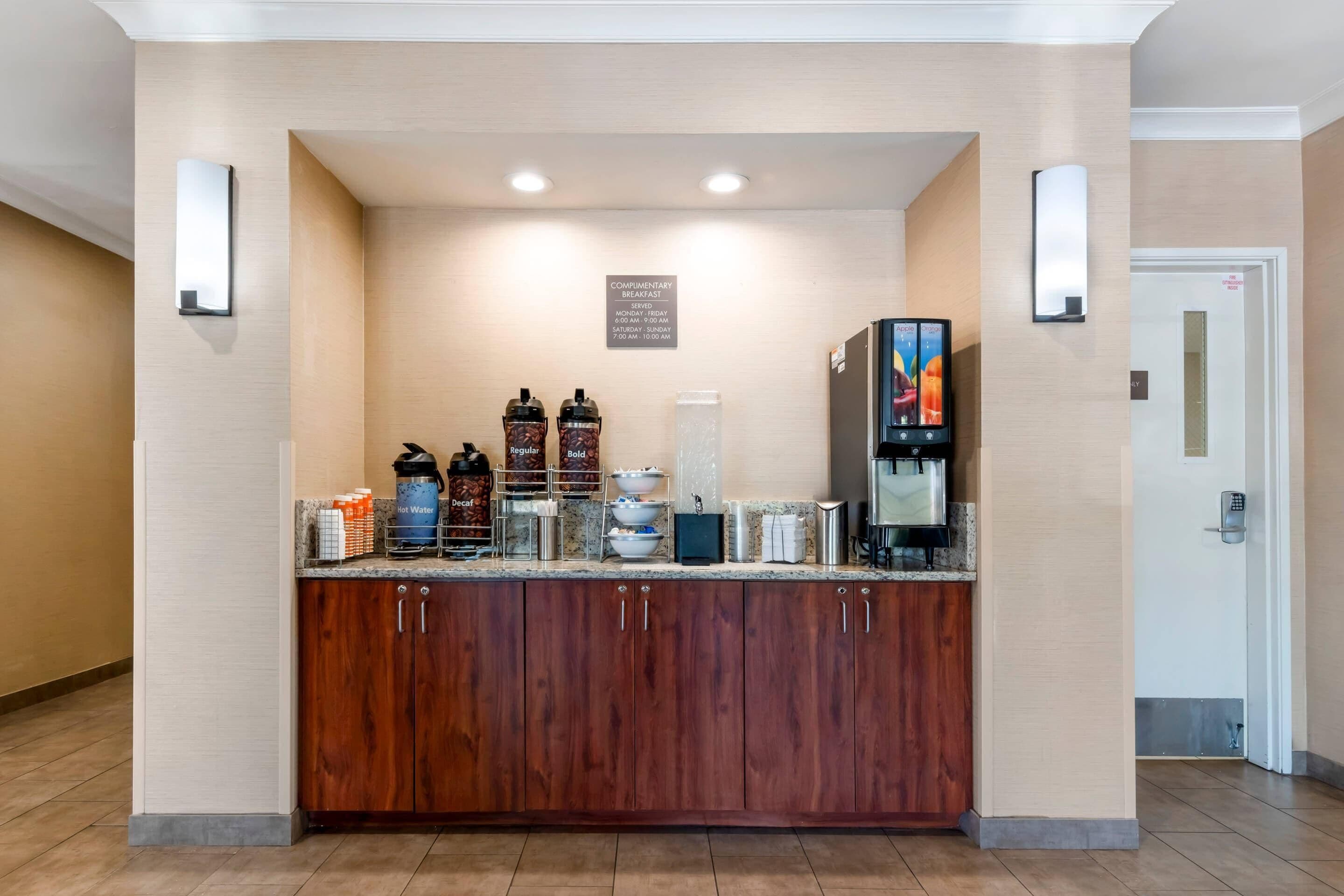 Zona de desayuno de hotel con mostrador de madera, máquinas de café y un cartel que indica los horarios de desayuno gratuito.