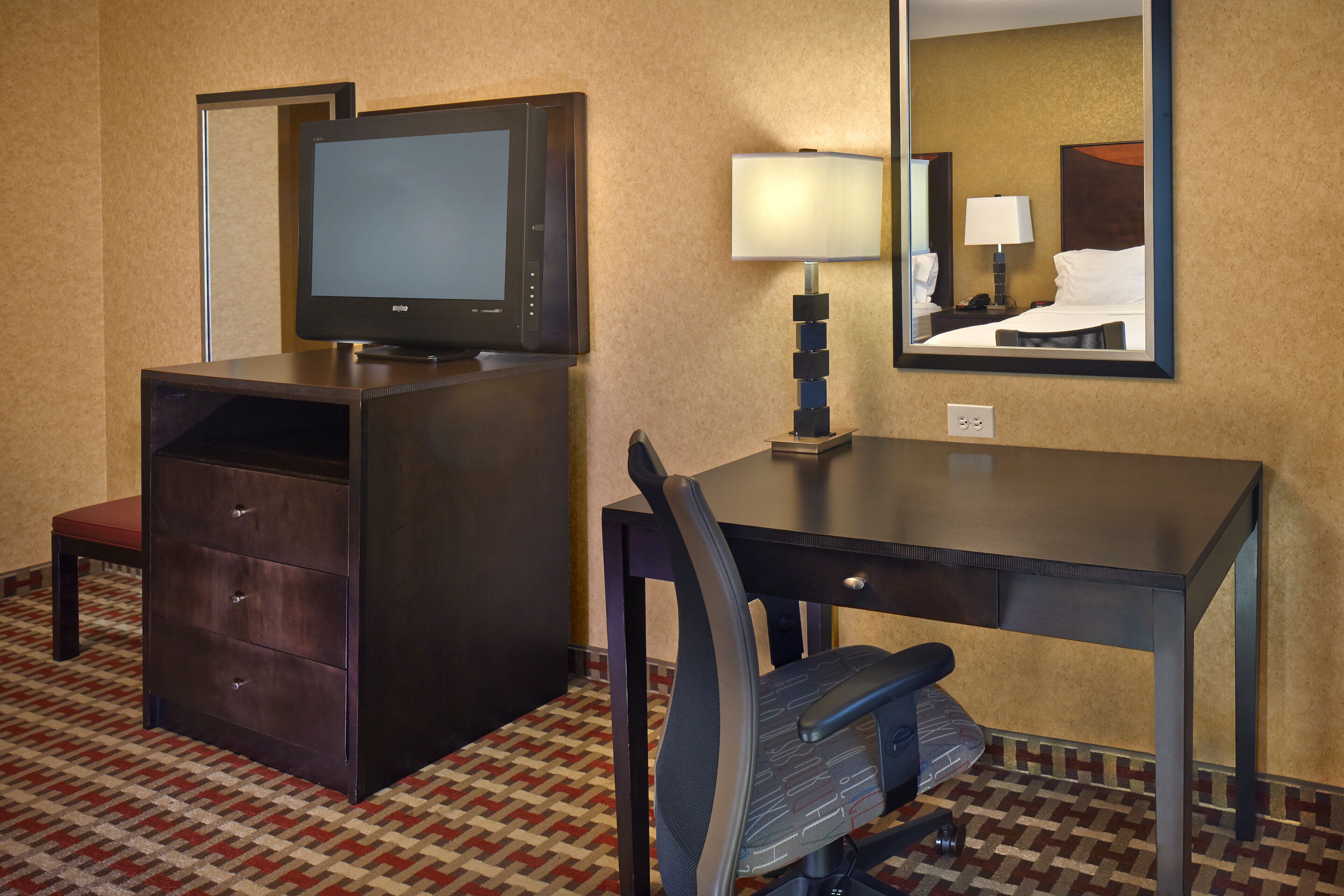 Une chambre d’hôtel équipée d’une télévision, d’un bureau, d’une chaise, d’une lampe et d’un miroir.
