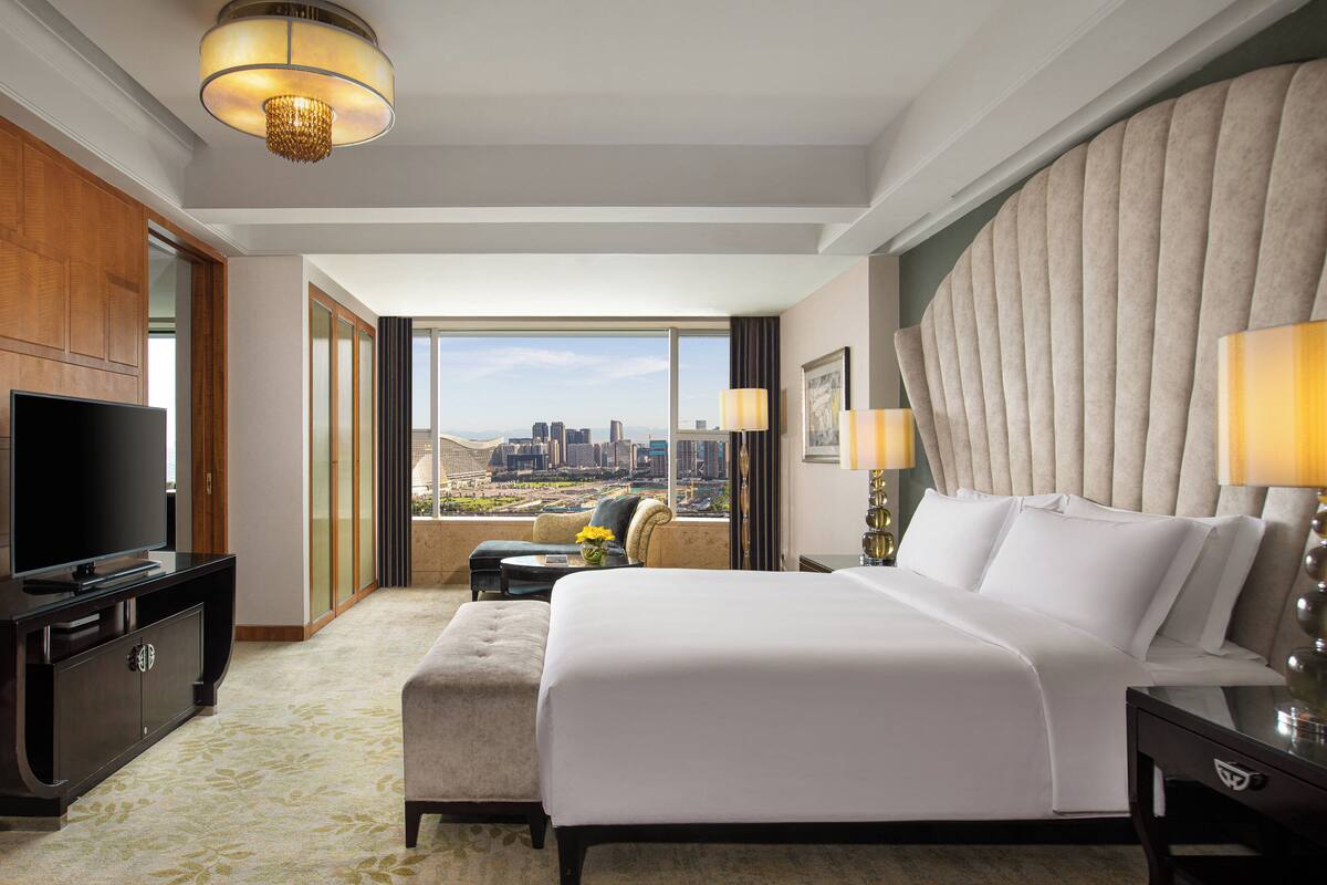 A hotel room with a large bed, a television, a desk, and a view of a cityscape.