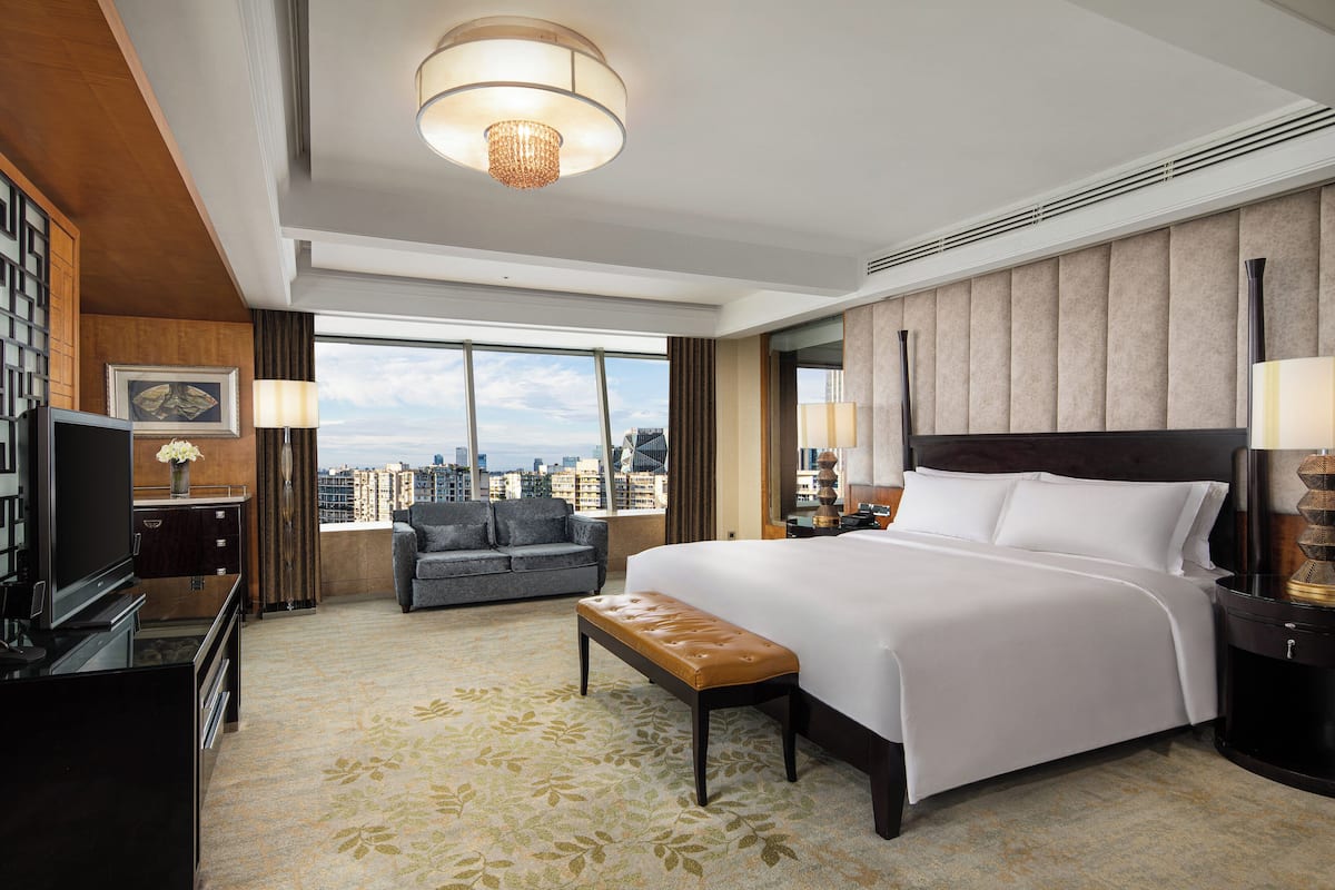 A hotel room with a large bed, a bench, a TV, a desk, and a view of the city.