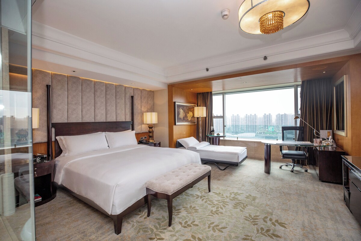 Habitación de hotel con una cama grande, un escritorio, una silla, un banco y vistas a la ciudad.