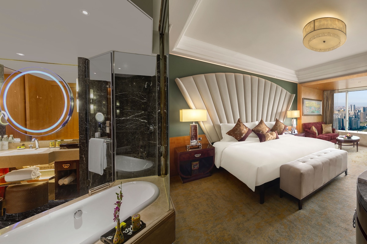 A modern hotel room with a large bed, a vanity with a mirror, a bathtub, and a city view.
