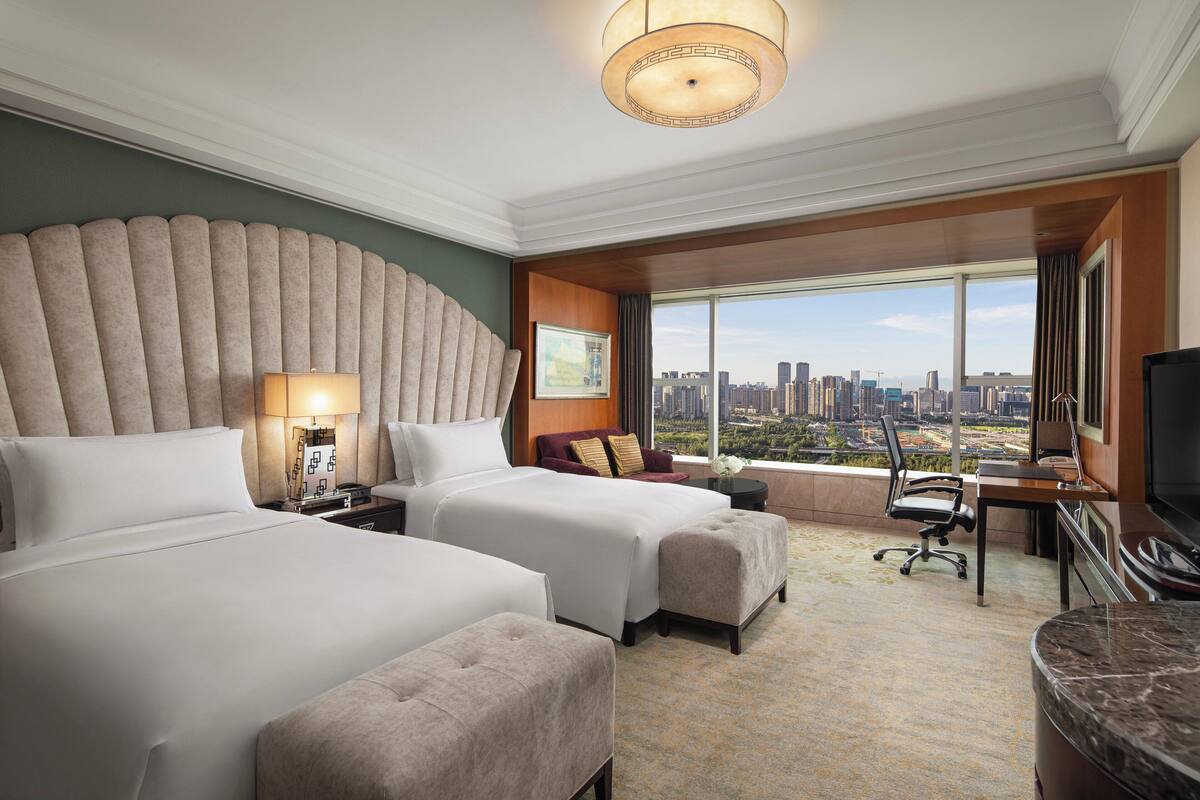 A hotel room with two beds, a desk, a chair, a TV, and a large window with a city view.