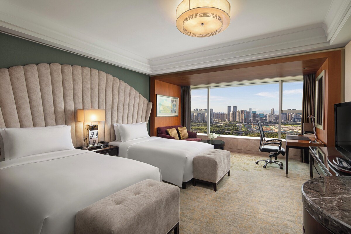 Habitación de hotel con dos camas, un escritorio, una silla, un televisor y un ventanal con vistas a la ciudad.