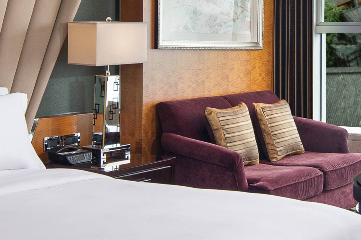 A hotel room with a bed, a desk with a lamp, a purple sofa with cushions, and a framed picture on the wall.