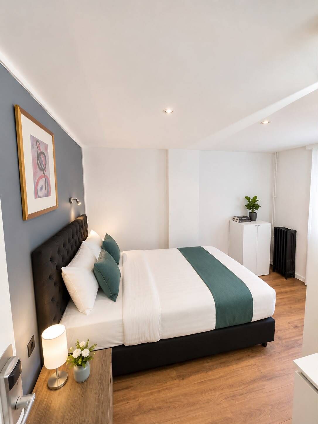 Une chambre d’hôtel moderne avec un grand lit, une table de chevet avec une lampe, un tableau encadré au mur et un radiateur dans un coin.
