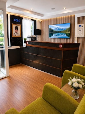 A reception desk with a digital clock, a television displaying a mountainous landscape, and a framed portrait.