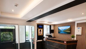 Un área de recepción moderna con un mostrador de madera, un reloj digital, un televisor y asientos verdes.